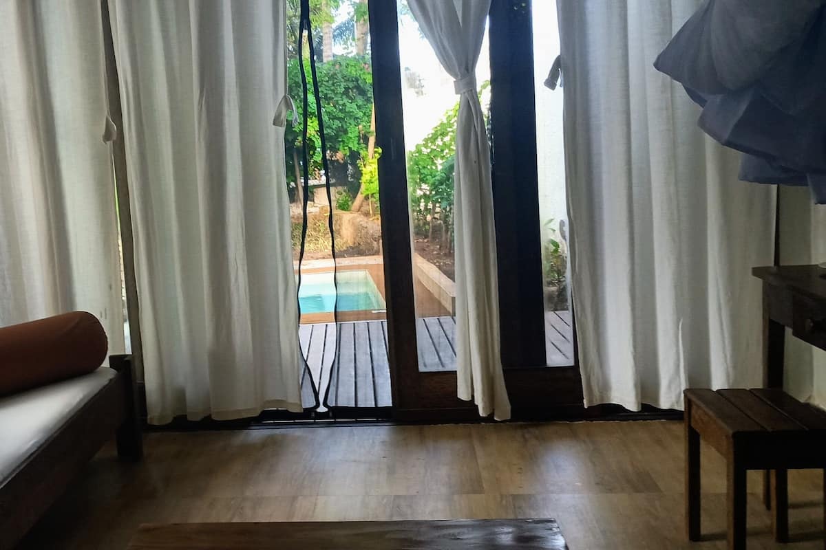 A room with a wooden floor, a bed with a brown pillow, a bench, and a view of a pool through a window with white curtains.