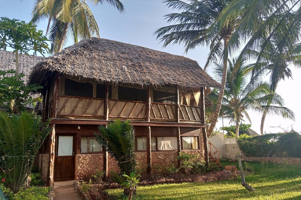 A two-story wooden house with a thatched roof, surrounded by lush greenery and palm trees.