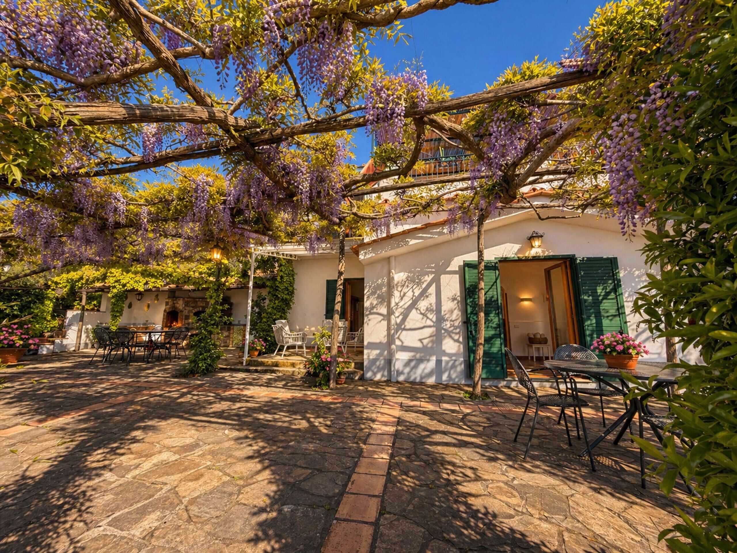 Een binnentuin met een pergola vol paarse bloemen, stenen vloer en buitenmeubilair.