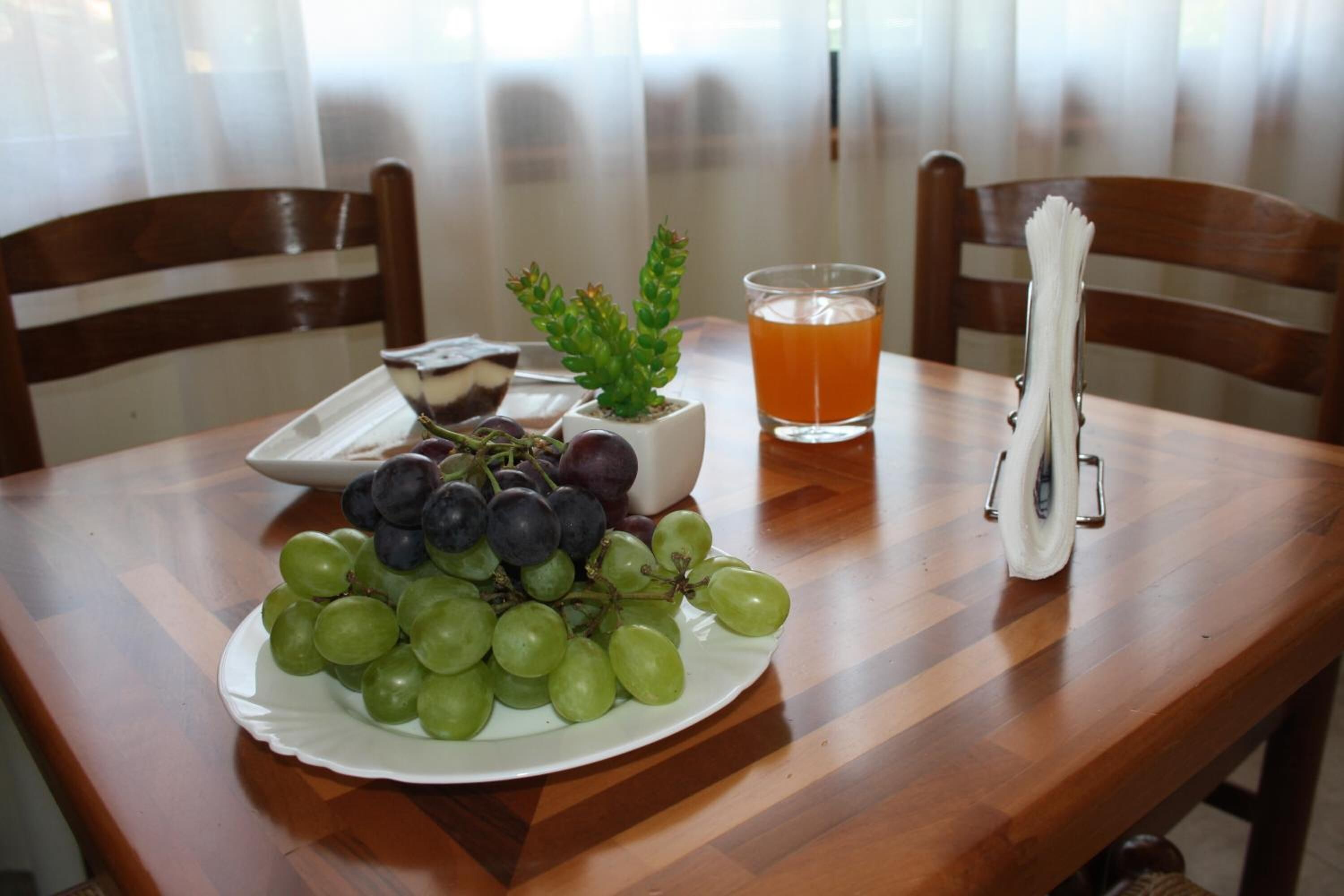 Una mesa de madera con un plato de uvas, una pequeña planta en maceta, un vaso de jugo de naranja y un portavasos.