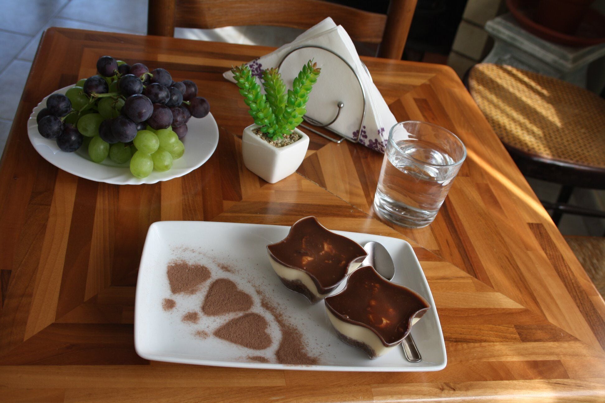 Una mesa de madera con una fuente de uvas, una pequeña planta en maceta, un vaso de agua y dos postres de chocolate.