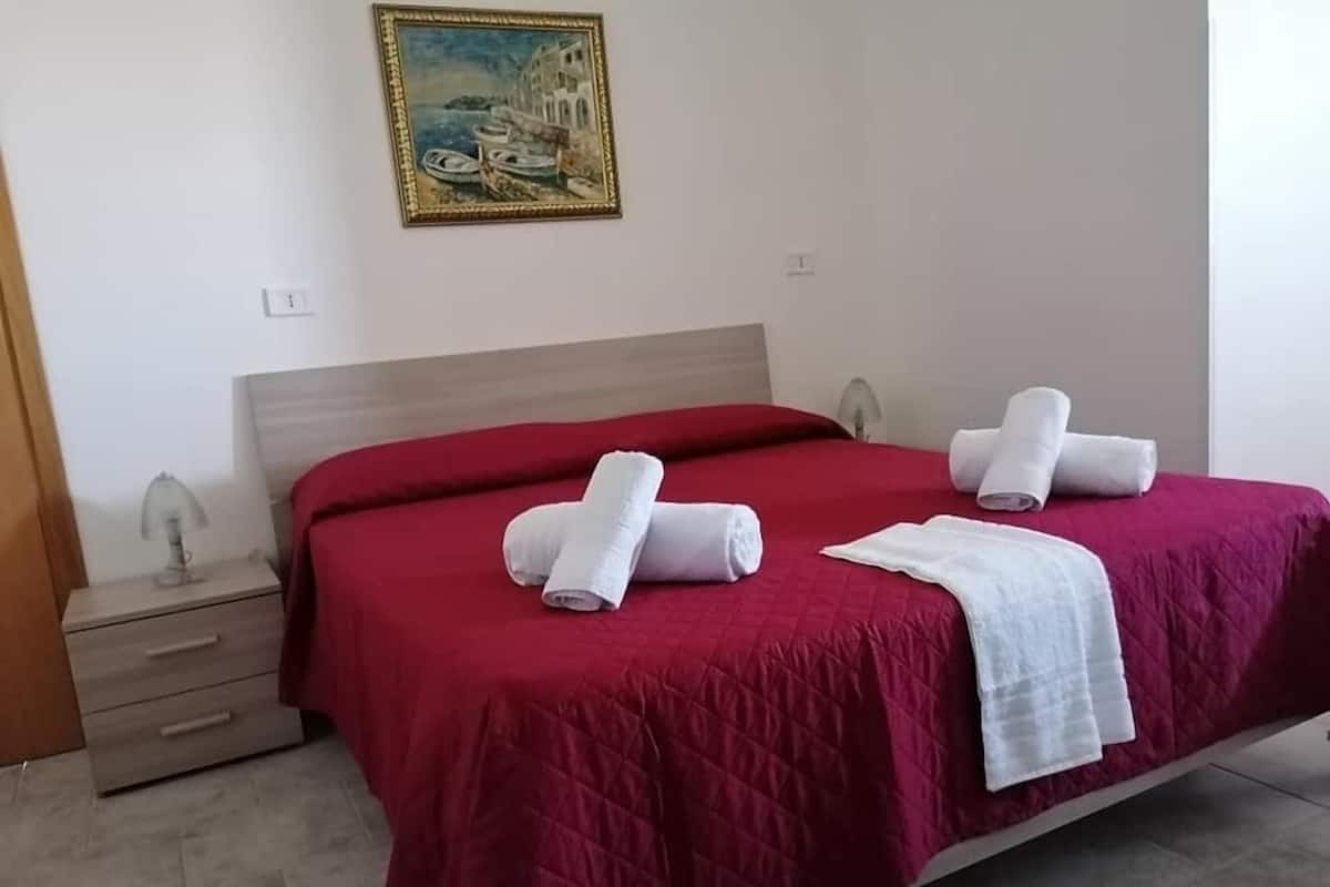 A neatly made bed with red bedding and white towels, a painting on the wall, and a wooden nightstand with a lamp.