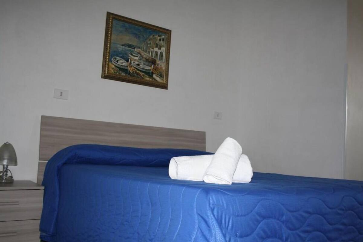 A hotel room with a bed, a painting on the wall, and a nightstand with a lamp.