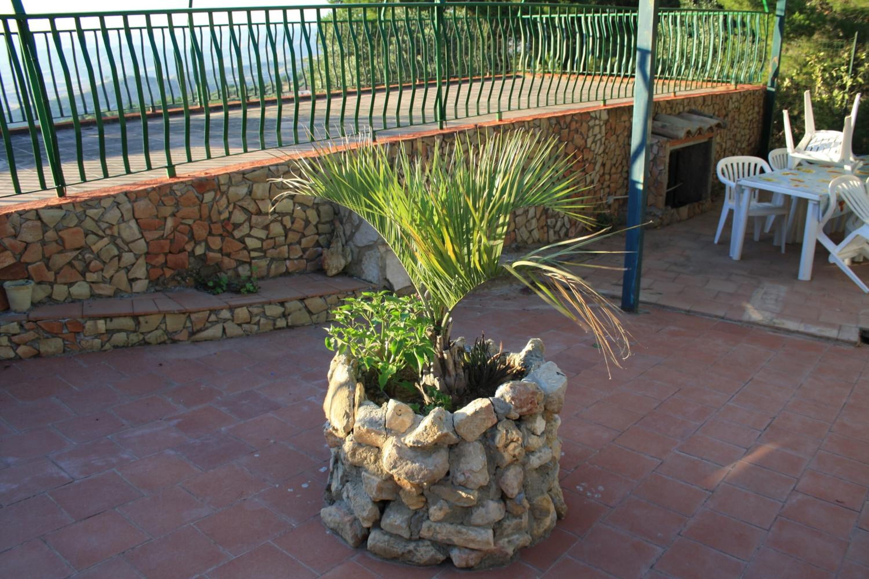 Ein Steinkasten mit Pflanze, eine Steinmauer und eine Terrasse mit weißen Stühlen und einem Tisch.