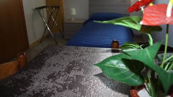 Une chambre avec une couvre-lit bleue, une chaise en bois, une petite table recouverte d’une nappe à motifs, et une plante en pot ornée de fleurs rouges.