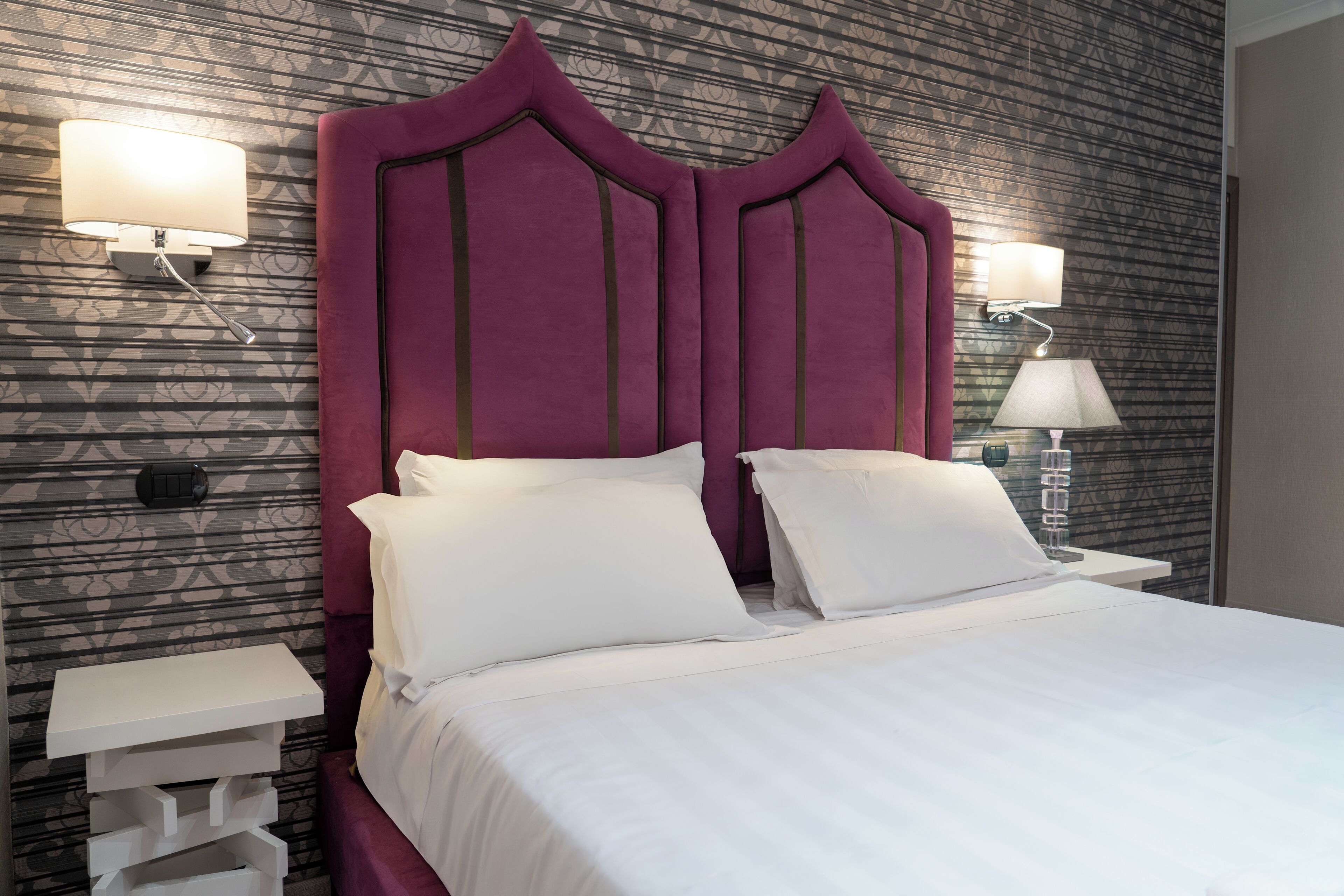 A neatly made bed with white linens, a purple upholstered headboard, and two bedside lamps.
