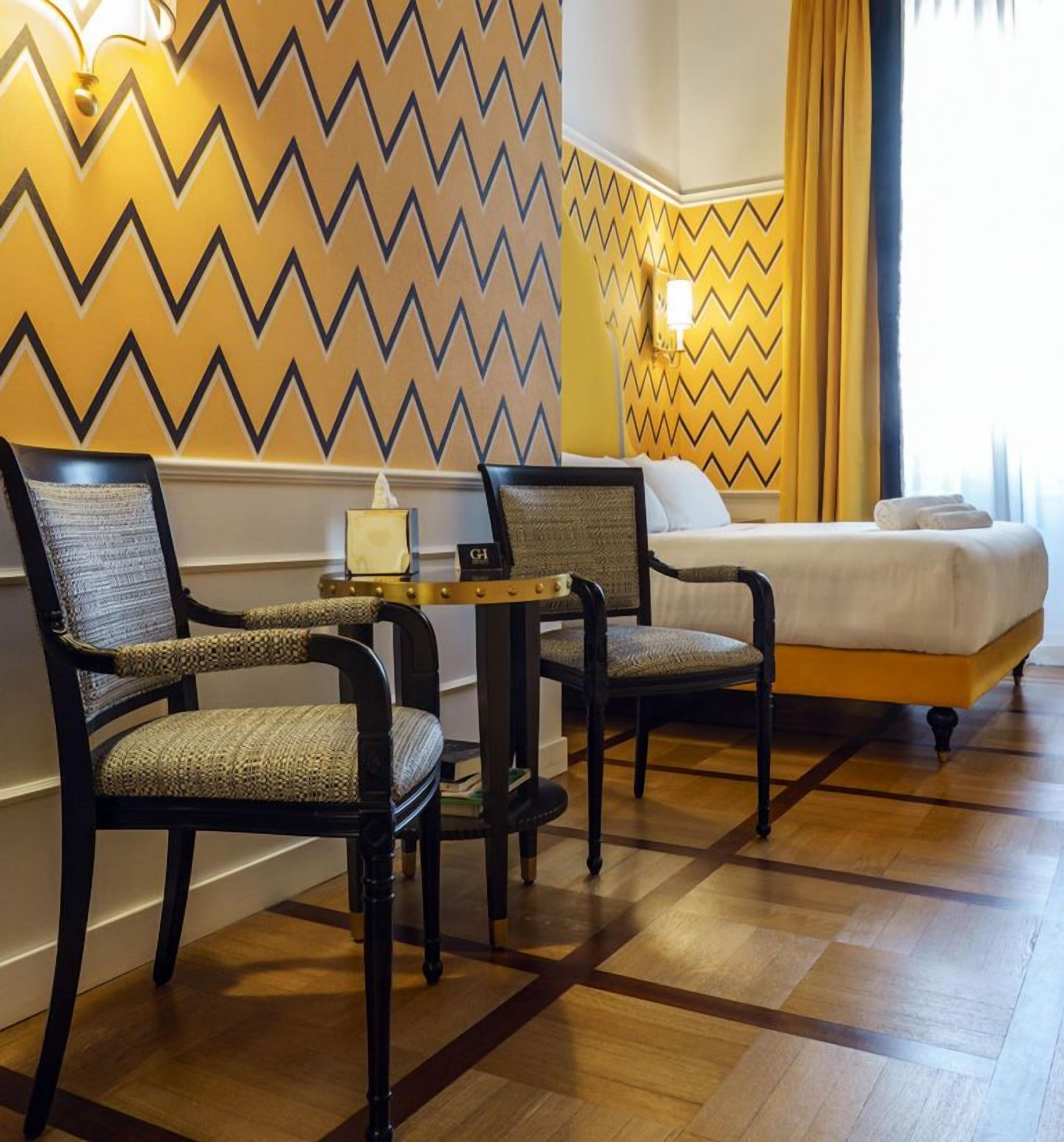 A hotel room with a bed, two armchairs, a small table with a lamp, and a wall with a zigzag pattern.