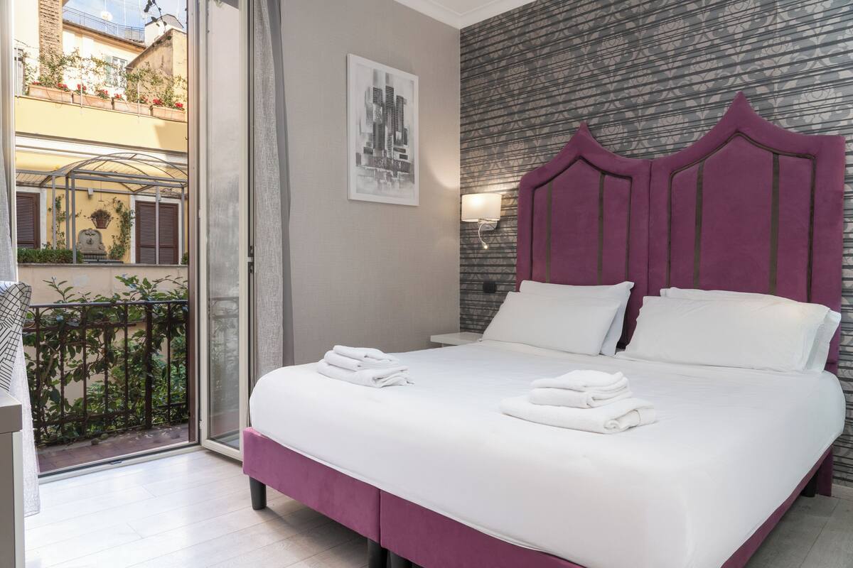 A hotel room with a bed, a view of the outside balcony, a cityscape painting, and a patterned wall.