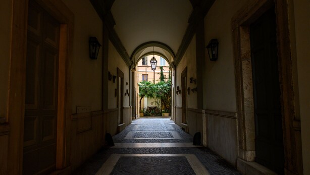 A corridor with a tiled floor, wooden doors, and a central archway leading to a courtyard with plants. - Il Campo Marzio (Rome)