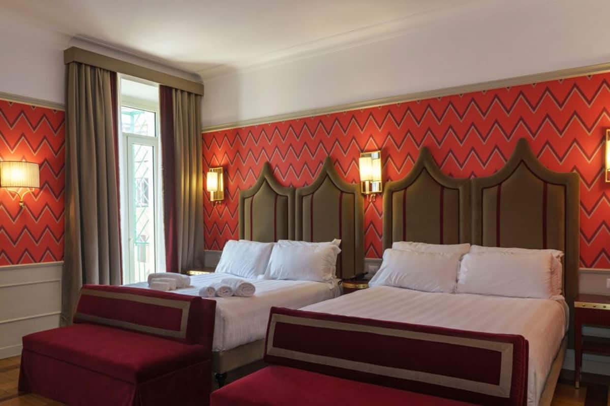 A hotel room with two beds, red upholstered benches, a large window with curtains, and a chevron-patterned wall.