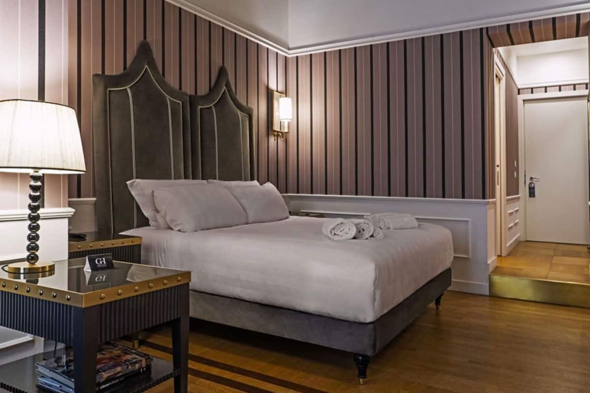 A hotel room with a bed, a bedside table with a lamp, a wall-mounted light, and a door.