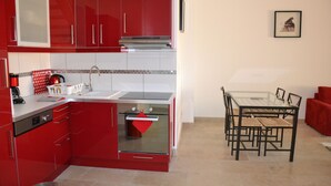 A compact kitchen with red cabinets, a built-in oven, and a dining area with a glass table and chairs.