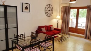 A living room with a red sofa, a glass coffee table, a black metal TV stand, a wall clock, and a window with curtains.