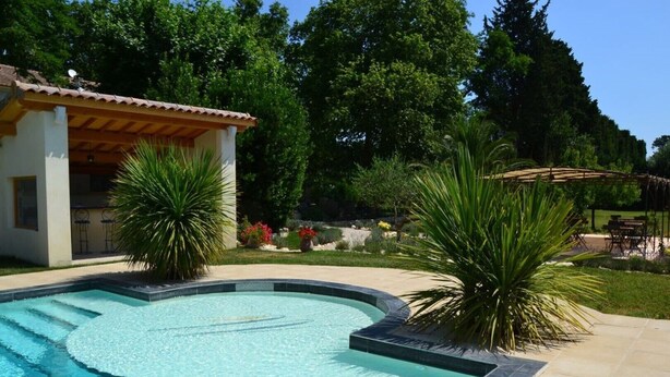 A swimming pool with a curved design, surrounded by a landscaped garden with palm trees and a patio area with outdoor furniture.