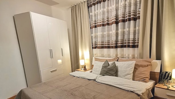 Ein ordentlich bezogenes Bett mit einem gemusterten Überwurf, ein Nachttisch aus Holz mit einer Lampe und ein Kleiderschrank mit Spiegel.