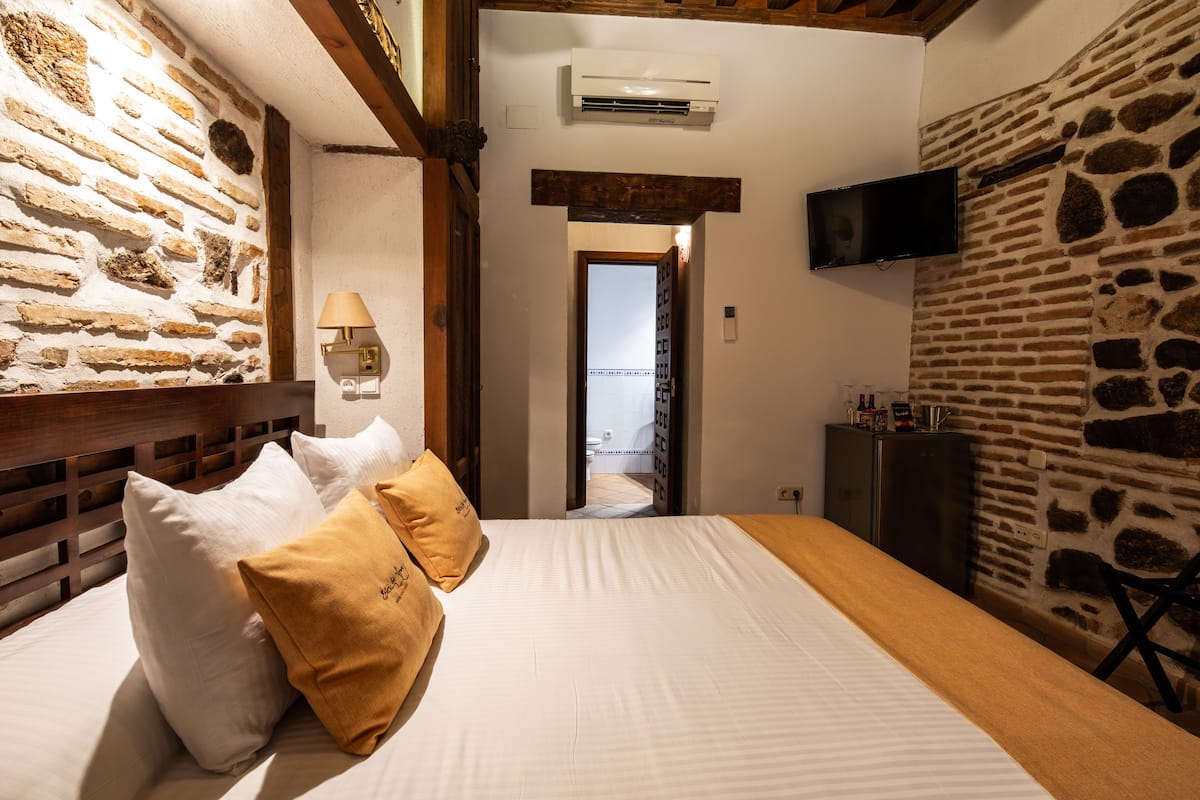 A hotel room with a stone wall, a bed with white and mustard pillows, a wooden headboard, a mounted TV, and a small cabinet.