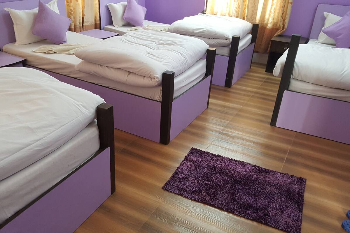 Ein Zimmer mit vier Einzelbetten, jeweils mit Holzrahmen und weißer Bettwäsche. Auf dem Holzboden liegt ein violetter Teppich, und auf der rechten Seite sind ein Paar blaue Hausschuhe zu sehen.