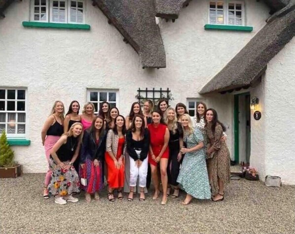 A group of people standing in front of a white building with green trim and a thatched roof.