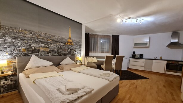 A modern bedroom with a large bed, a cityscape mural, a dining area, and a kitchenette.