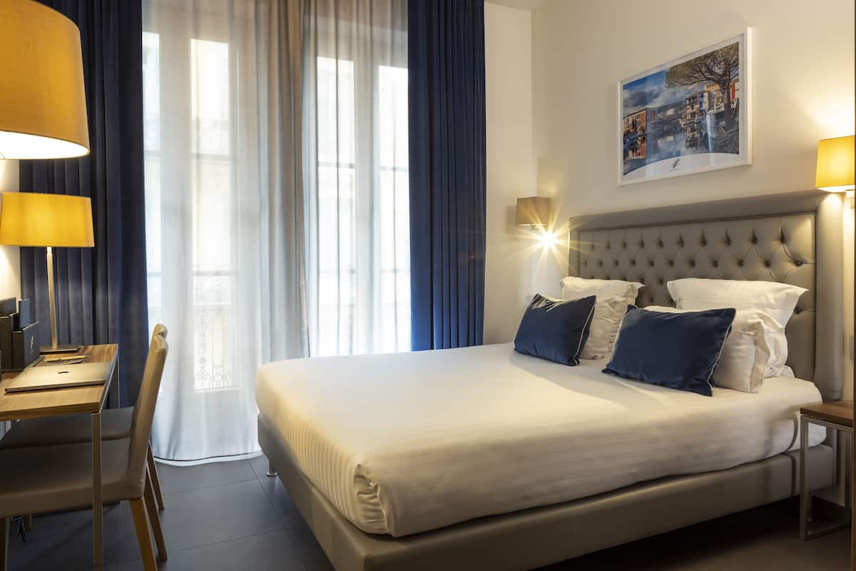 A hotel room with a large bed, a desk, and a chair. There is a window with curtains, a wall-mounted lamp, and a framed picture on the wall.