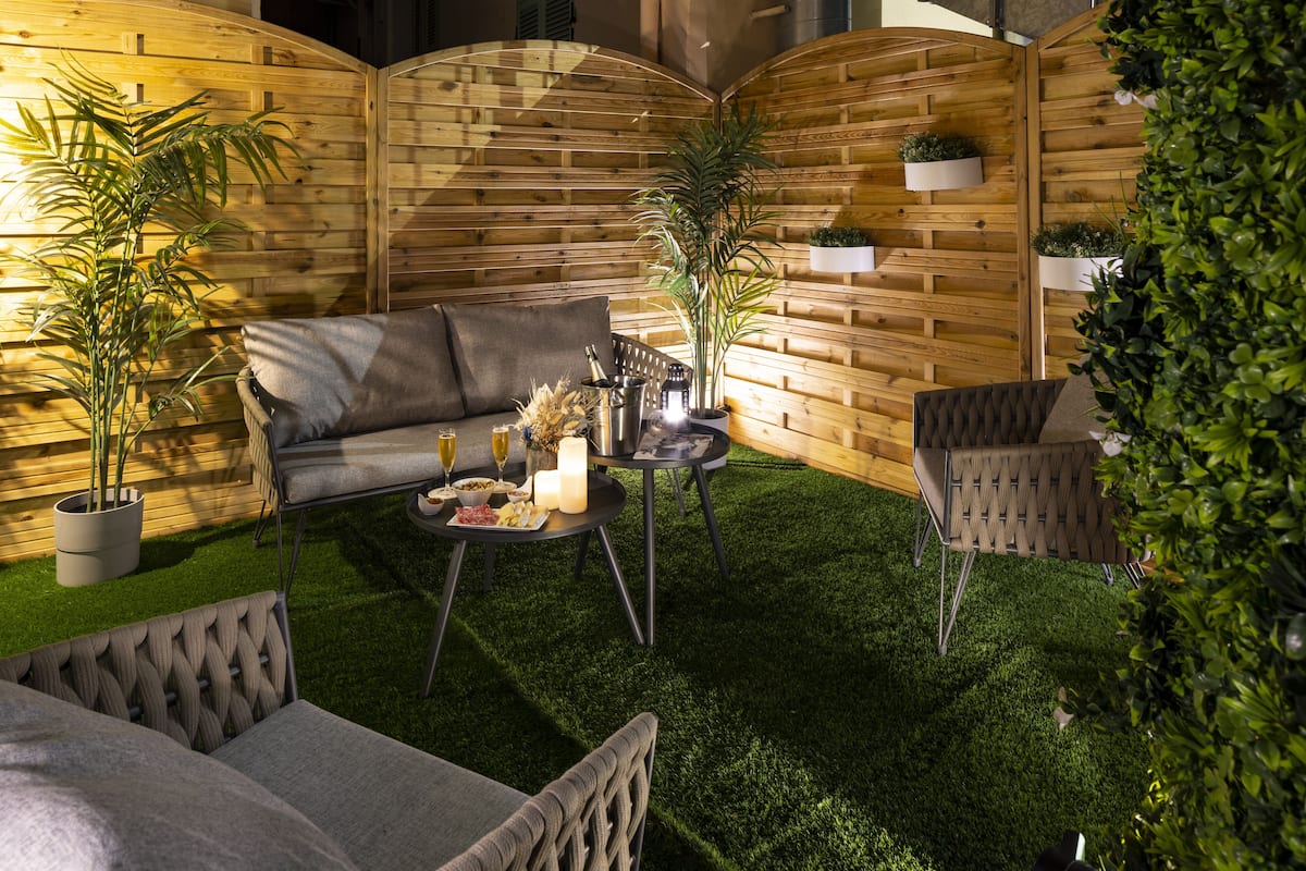 An outdoor patio with a wooden wall, a sofa, a dining table with candles, and potted plants.