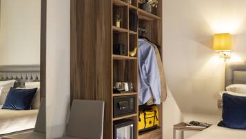 A hotel room with a bed, a seating area with a chair, a built-in wardrobe with shelves, a bedside table with a lamp, and a small refrigerator.