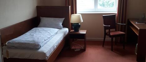 A hotel room with a single bed, a desk, a chair, and a window with curtains.