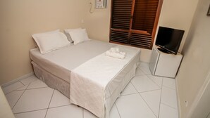 A neatly made bed with a white bedspread and pillows, a bedside table with a television, and a window with wooden blinds. - Suites Natalia Enseada (Guarujá)