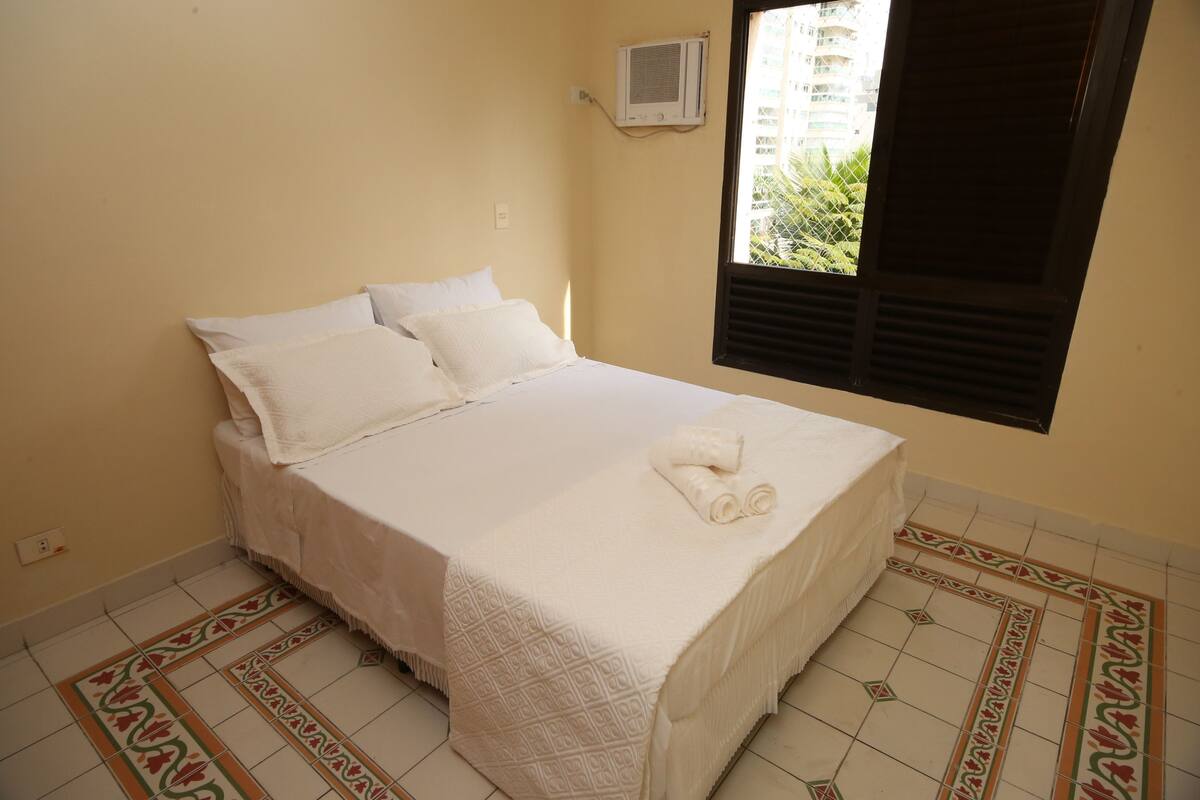 A neatly made bed with white linens and a rolled towel on top, a window with blinds, and a patterned floor.