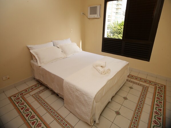 A neatly made bed with white linens and a rolled towel on top, a window with blinds, and a patterned floor. - Suites Natalia Enseada (Guarujá)