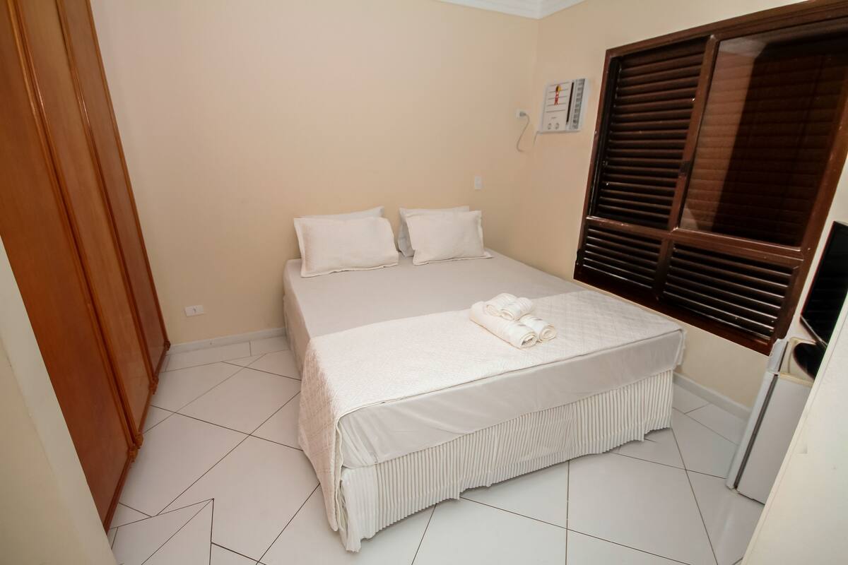 A bedroom with a bed, pillows, a window with blinds, and a thermostat on the wall.