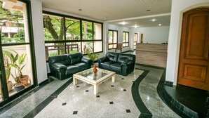 A spacious lobby with leather sofas, a glass coffee table, and large windows offering views of greenery. - Suites Natalia Enseada (Guarujá)