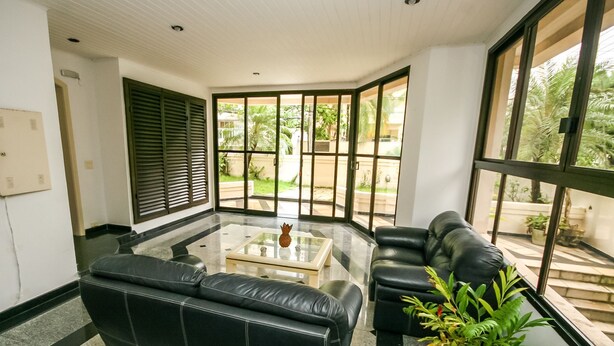 A modern living room with a black leather sofa, a glass coffee table, and a potted plant. - Suites Natalia Enseada (Guarujá)
