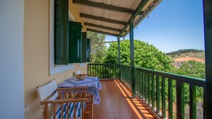 En balkon med bord og stole, et grønt gelænder og udsigt til grøn vegetation og bakker.
