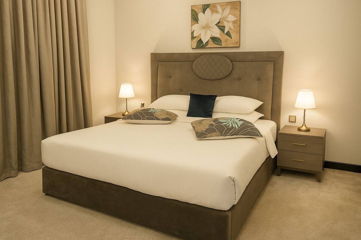 A hotel room with a large bed, two bedside tables with lamps, a headboard with a decorative panel, and a painting above the bed.