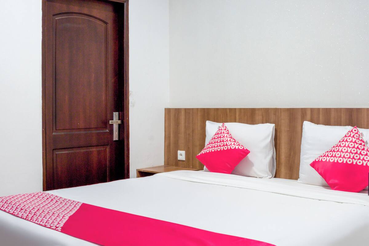 Hotellihuone, jossa on puinen ovi, valkoisilla lakanoiden ja kahdella vaaleanpunaisella tyynyllä varustettu sänky sekä yöpöytä, jossa on lamppu.