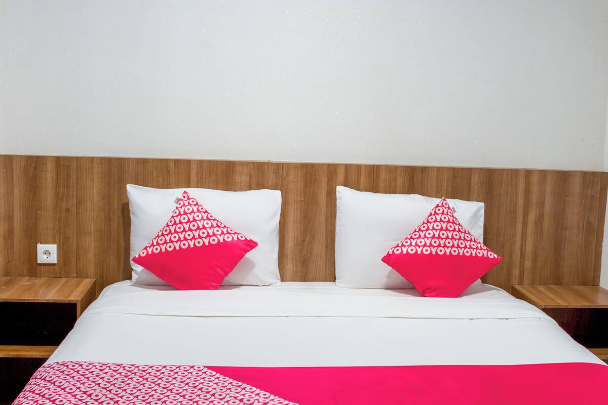 Hotellihuone, jossa on puinen sängynpääty, kaksi valkoista tyynyä, joissa on punainen teksti, valkoinen päiväpeite ja punavalkoinen kuviollinen peitto.