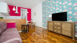 A modern living room with a herringbone wooden floor, a pink sofa, a wooden TV stand, and a floral wallpaper. - Crown Apartment (Zagreb)