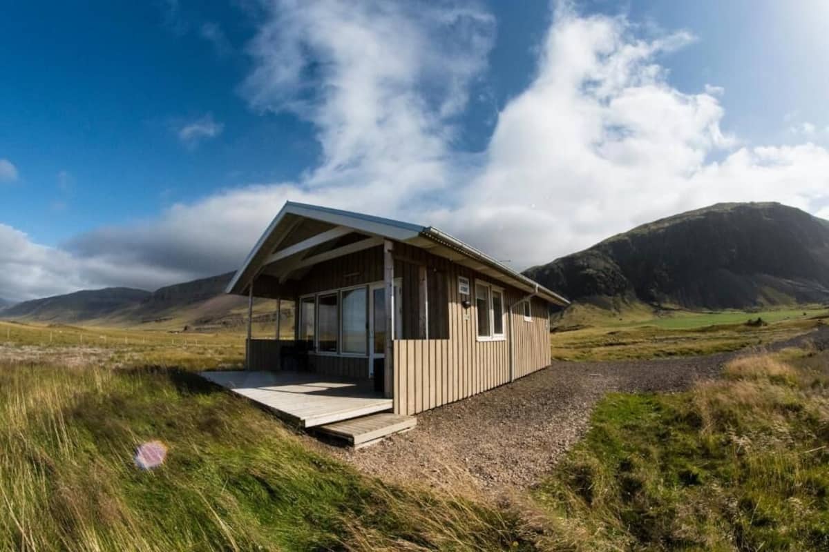 Una cabaña de madera con fachada de vidrio, ubicada en un campo cubierto de hierba con montañas al fondo.