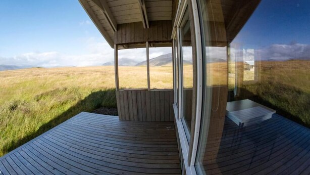 Une terrasse en bois, dotée d’une paroi vitrée, qui offre une vue sur un champ verdoyant et des montagnes lointaines.
