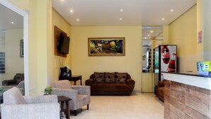 Un área de recepción con un sofá marrón, dos sillones, un televisor montado en la pared y un refrigerador Coca-Cola.