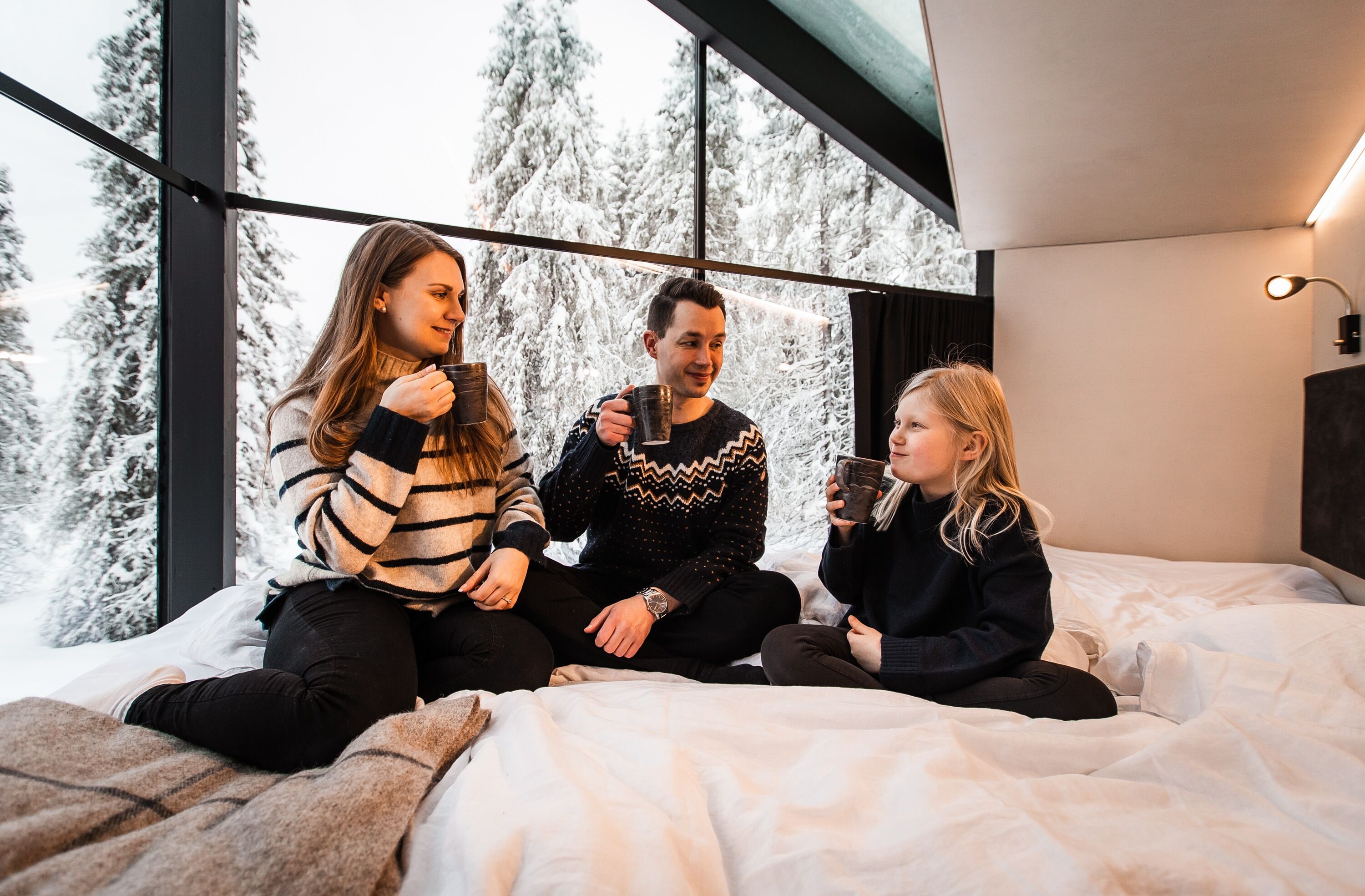 Tres personas sentadas en una cama, con una ventana grande detrás y una lámpara en la pared, frente a una vista de árboles cubiertos de nieve.