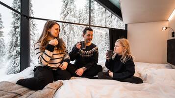 Tres personas sentadas en una cama, con una ventana grande detrás y una lámpara en la pared, frente a una vista de árboles cubiertos de nieve.