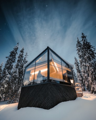 A glass-enclosed cabin with a wooden base, surrounded by snow-covered trees, featuring a warm interior light.