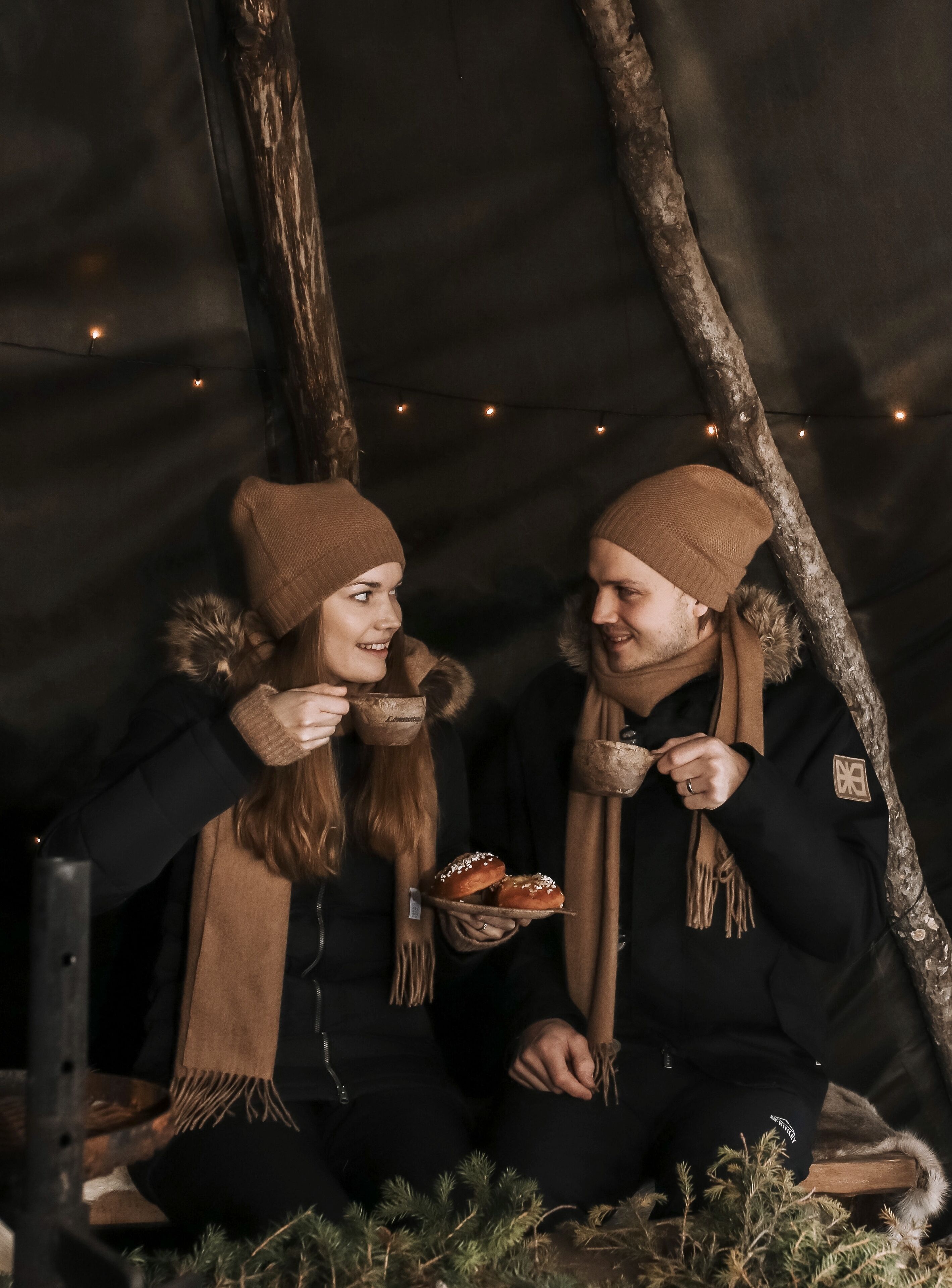 Two people sitting on a bench in a tent-like structure with string lights, holding pastries.