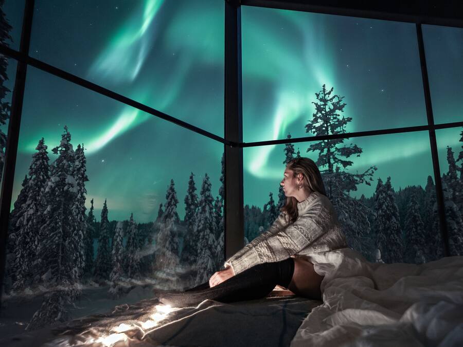 Una persona sentada en una cama en una habitación con ventanas amplias, observando un bosque nevado y las auroras boreales.