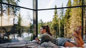 A person lying on a bed in a room with large windows overlooking a forest and a body of water. - Magical Pond Nature Igloos (Rukatunturi)