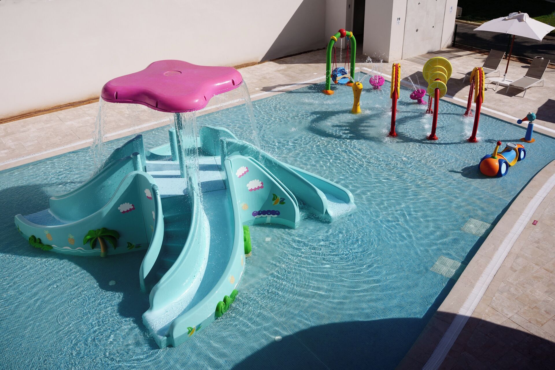 Une piscine dotée de plusieurs toboggans et d’installations ludiques aquatiques, notamment un siège rose et divers jouets aquatiques.