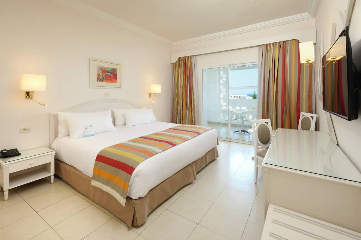 Une chambre d’hôtel avec un grand lit, un bureau, une chaise, une télévision et un balcon avec des rideaux.