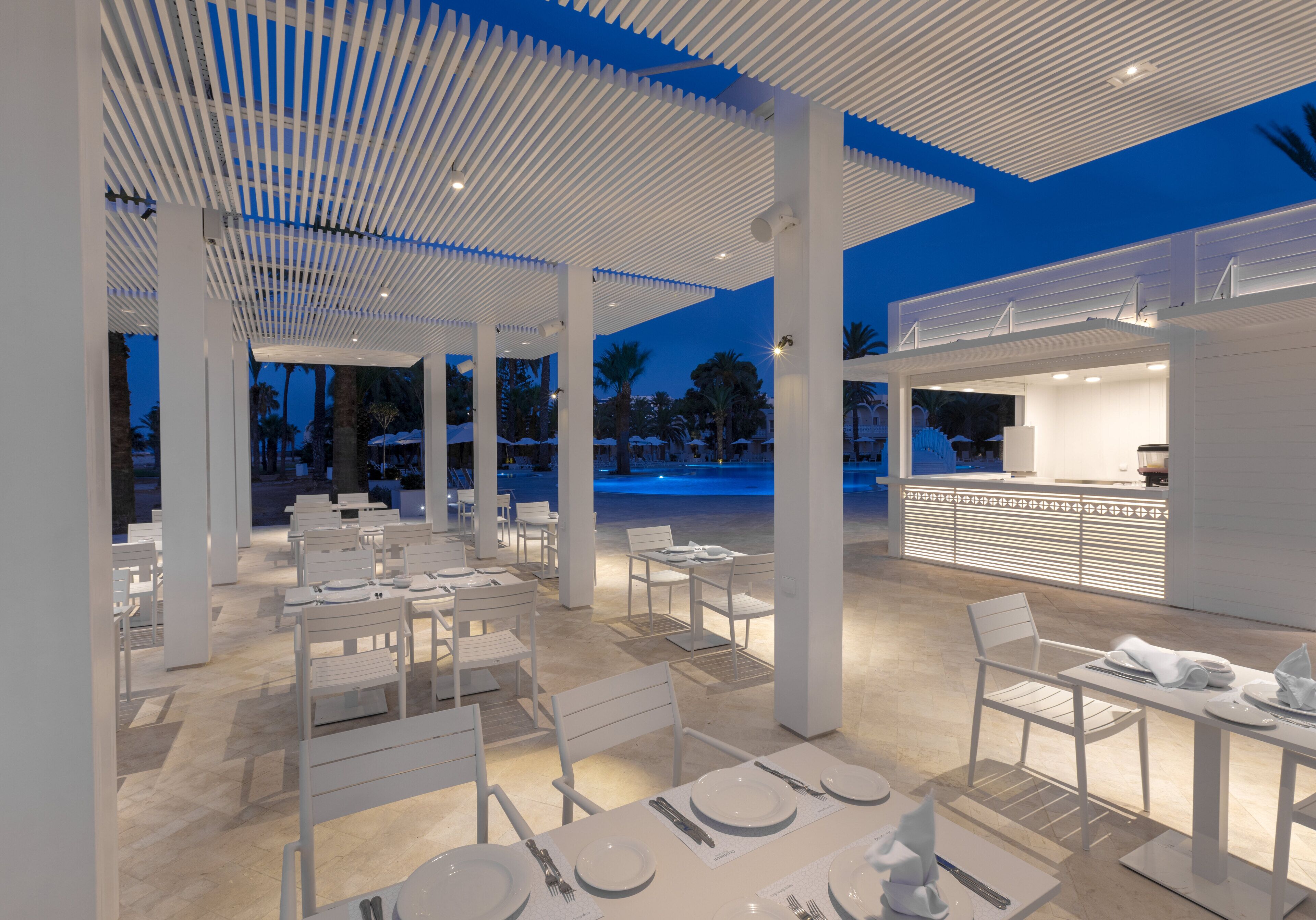A spacious outdoor dining area with white furniture and a modern design, featuring a bar and seating with a view of the surrounding landscape.